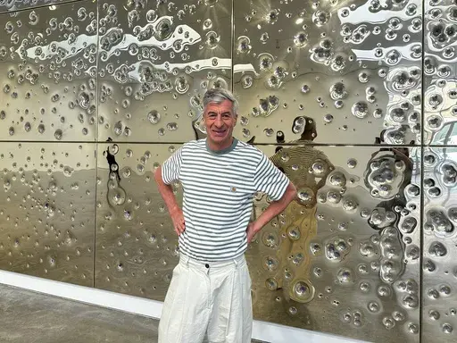 Italian artist Maurizio Cattelan poses with his installation, "Sunday," at Gagosian gallery on May 1, 2024, in New York. The installation consists of 64 panels plated with 24-karat gold and pockmarked by gunfire. (AP Photo/Jocelyn Noveck)