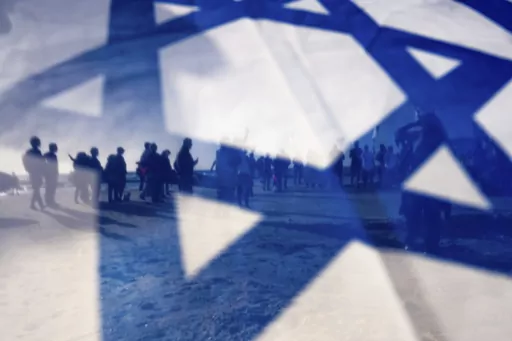Israeli demonstrators are seen through Israel's national flag during a protest against plans by Prime Minister Benjamin Netanyahu's government to overhaul the judicial system, in Cesarea, Israel, Friday, March 17, 2023. (AP Photo/Oded Balilty)