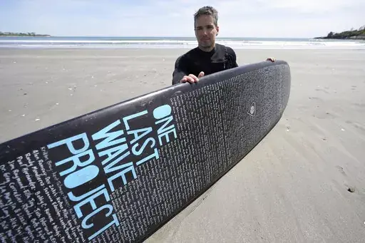 Dan Fischer, of Newport, R.I., sits for a photograph with his surfboard on Easton's Beach, in Newport, Wednesday, May 18, 2022. Fischer, 42, created the One Last Wave Project in January 2022 to use the healing power of the ocean to help families coping with a loss, as it helped him following the death of his father. Fischer places names onto his surfboards, then takes the surfboards out into the ocean as a way to memorialize the lost loved ones in a place that was meaningful to them. (AP Photo