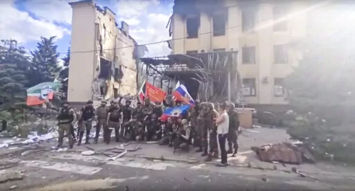 This photo taken from video provided by Ramzan Kadyrov's Official Telegram channel released on Saturday, July 2, 2022, shows Russian troops including soldiers of Chechen regiment waving Russian and Chechen republic national flags as they pose for a photo in front of a destroyed building in Lysychansk, Ukraine. After more than four months of ferocious fighting, Russia claimed full control over one of the two provinces in Ukraine’s eastern industrial heartland. (Ramzan Kadyrov's Official Telegra