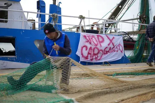 A fisherman mends a net in front of his fishing boat with a banner reading "stop for gasoline increase", in the Roman port of Fiumicino, Friday, March 11, 2022. Fishermen, facing huge spikes in oil prices, stayed in port, mending nets instead of casting them. Nowhere more than in Italy, the European Union’s third-largest economy, is dependence on Russian energy taking a higher toll on industry. (AP Photo/Andrew Medichini)