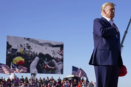 As footage from the Jan. 6, 2021, insurrection at the U.S. Capitol is displayed in the background, former President Donald Trump stands while a song, "Justice for All," is played during a campaign rally at Waco Regional Airport, Saturday, March 25, 2023, in Waco, Texas. The tune, “Justice for All,” is the Star-Spangled Banner and it was sung by a group of defendants jailed over their alleged roles in the January 2021 insurrection. The national anthem is overlaid with Trump reciting the Pledg