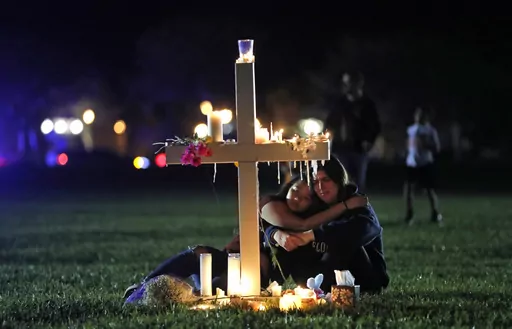 People comfort each other as they sit and mourn at one of seventeen crosses, Feb. 15, 2018, after a candlelight vigil for the victims of the shooting at Marjory Stoneman Douglas High School, in Parkland, Fla. A reenactment of the 2018 massacre that left 17 dead, 17 wounded and hundreds emotionally traumatized, is scheduled to be conducted Friday, Aug. 4, 2023, as part of lawsuits filed by the victims' families and the injured. (AP Photo/Gerald Herbert, File)