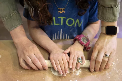 Wearing a necklace with her name in Hebrew, Charlotte Gleicher, 7, a first grader at Milton Gottesman Jewish Day School of the Nation's Capital, is helped by her teacher, Dafna Kiverstein, to roll out dough for matzah during a "Matzah Factory" field trip at the JCrafts Center for Jewish Life and Tradition in Rockville, Md., Thursday, April 18, 2024, ahead of the Passover holiday. To be kosher for Passover, which begins next Monday evening, the dough has to be prepared and cooked all within 18 mi