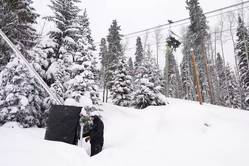 Ian Sidwell adjusts a machine used to make snow at Vail Mountain Resort as snowboarders ride a lift, Wednesday, Dec. 29, 2021, in Vail, Colo. Newer snowmaking technology is allowing ski areas to be more efficient with energy and water usage as climate change continues to threaten snowpack levels. (AP Photo/Brittany Peterson)